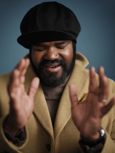 Close-up studio portrait of a bearded African-American man in a coat and cap with hands raised, against a solid blue backdrop.