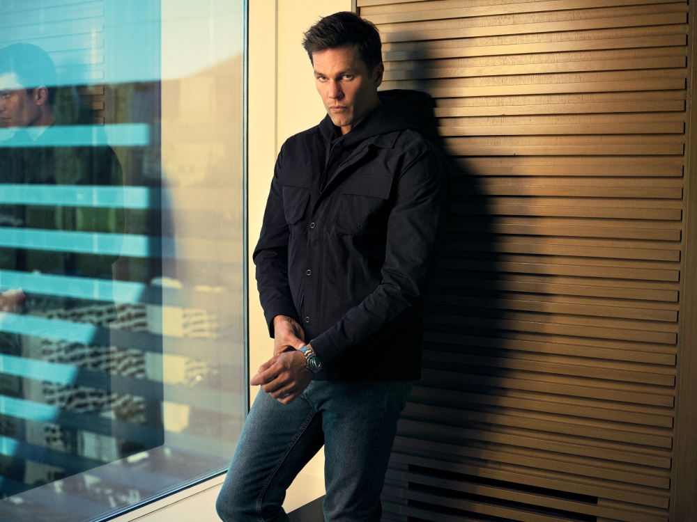 Portrait of an adult male in a black jacket and jeans posing next to a window with natural lighting and a wood-panelled wall.