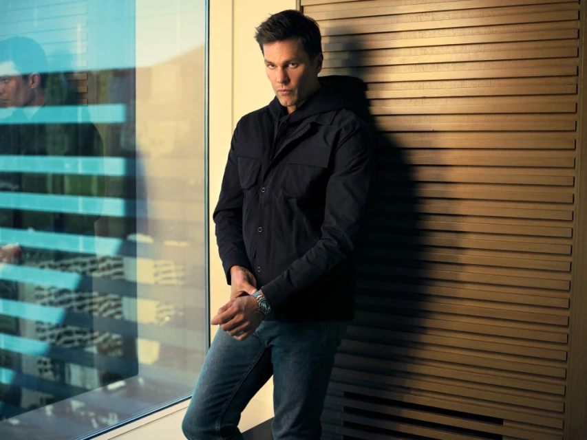 Portrait of an adult male in a black jacket and jeans posing next to a window with natural lighting and a wood-panelled wall.