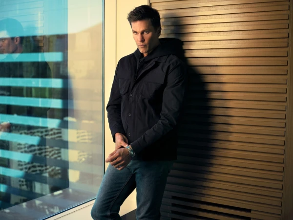 Portrait of an adult male in a black jacket and jeans posing next to a window with natural lighting and a wood-panelled wall.