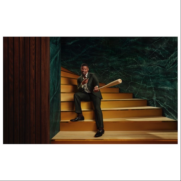 Well-dressed adult male sitting on stairs with a baseball bat, against a marble and wood panelled wall in a neutral indoor setting.