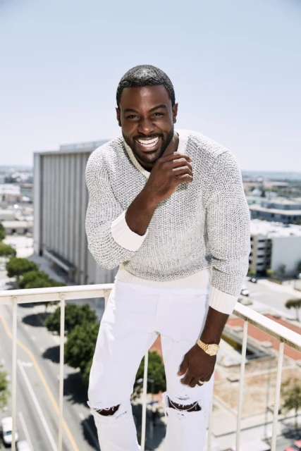 Smiling African-American man posing outdoors on a sunny day wearing a grey sweater and white jeans with a city building backdrop.