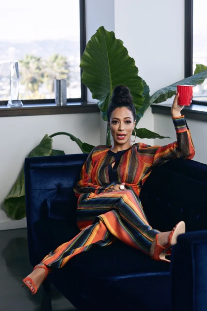 Woman in a striped suit sits in a blue chair, holding a red cup, with an interior view of a city and foliage behind her.