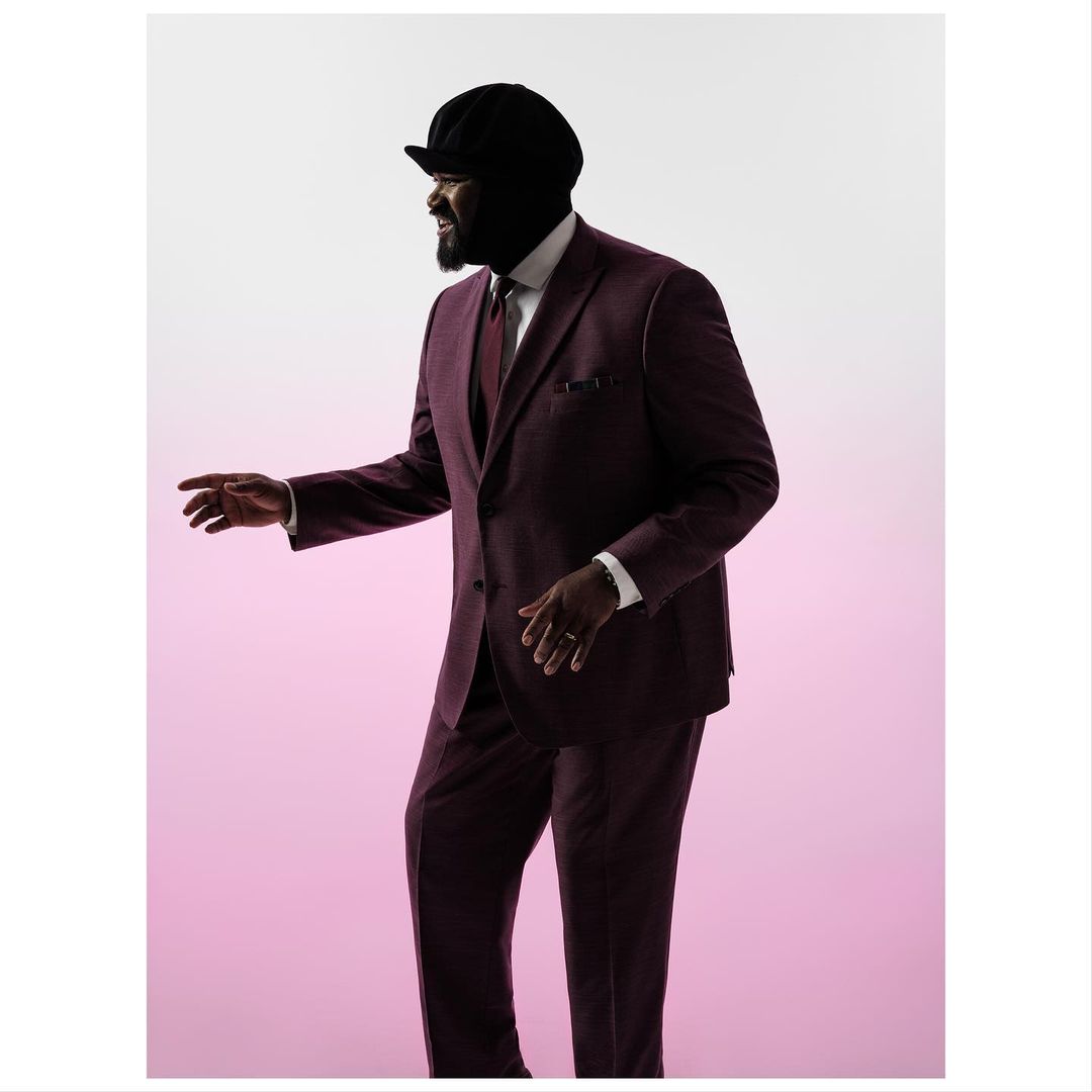A stylish African American man in a burgundy suit and cap, looking away and slightly smiling against a pink gradient background.