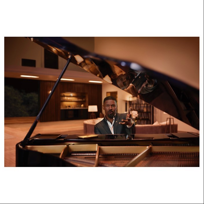 Adult male playing a grand piano in a modern living room. The piano is black, and the man is wearing a grey suit.
