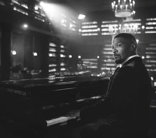 Black and white portrait of an African-American man playing a grand piano in an ornate room with glowing chandeliers.