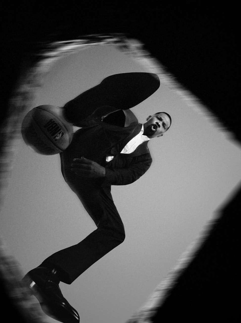 A man in a suit dribbles a basketball from a low angle, creating a dynamic black-and-white image with a blurred background.