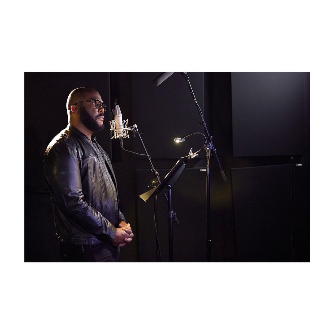 Adult man in a leather jacket singing into a microphone in a dimly lit music studio, side view.