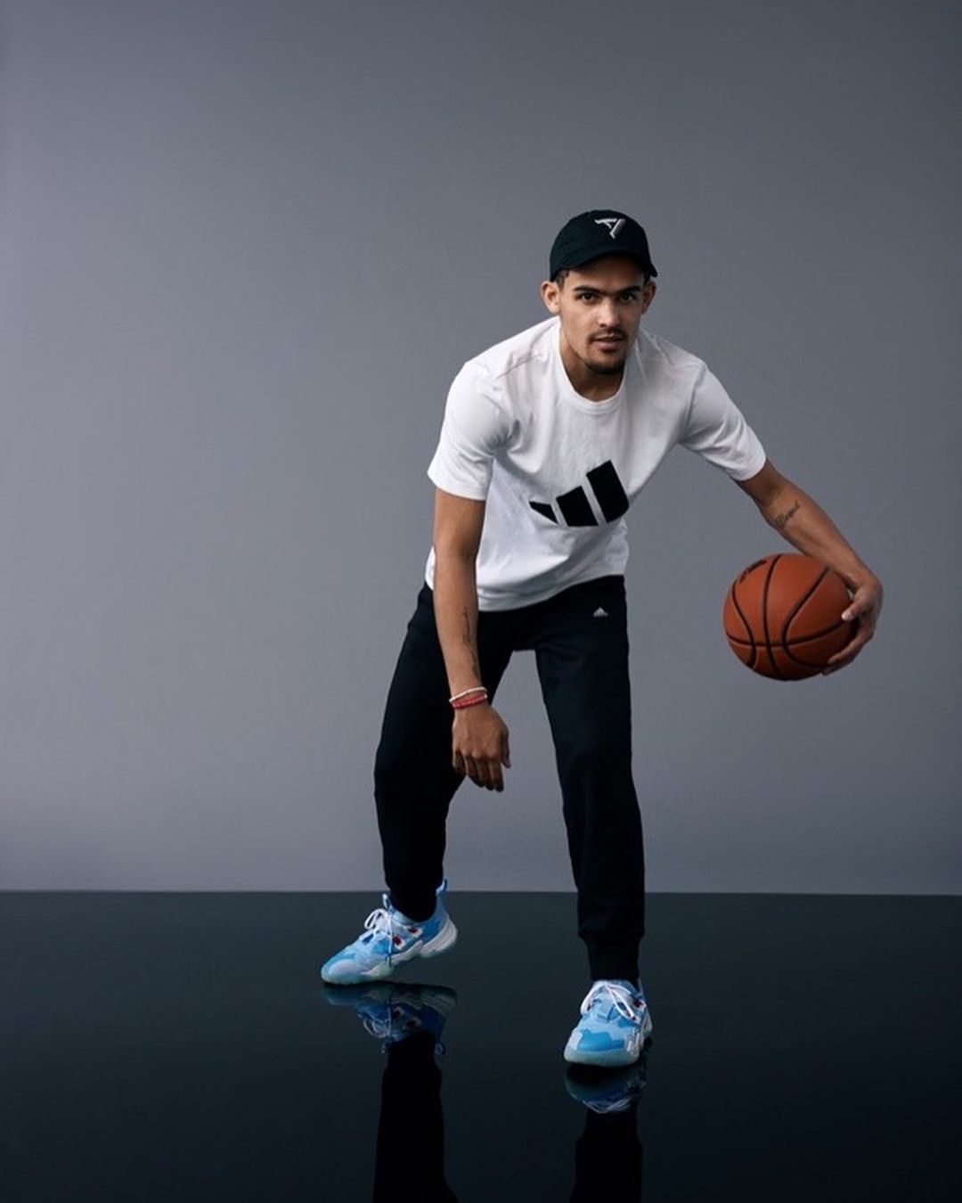 Adult male poses in a photo studio, wearing athletic wear and dribbling a basketball on a reflective floor against a gray background.