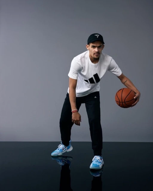 Adult male poses in a photo studio, wearing athletic wear and dribbling a basketball on a reflective floor against a gray background.