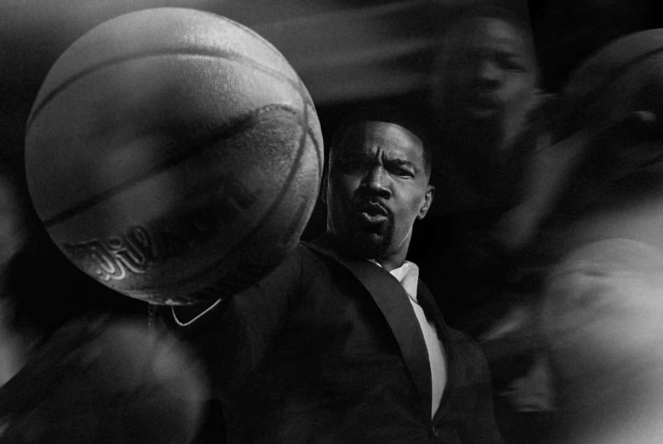Monochromatic portrait of an adult male in a suit holding a basketball, with multiple blurred human figures in the background.