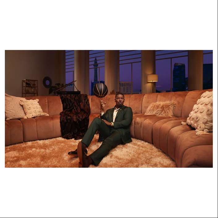 Man in green suit sits on a large couch and spins a basketball on his finger in an apartment with a cityscape view.
