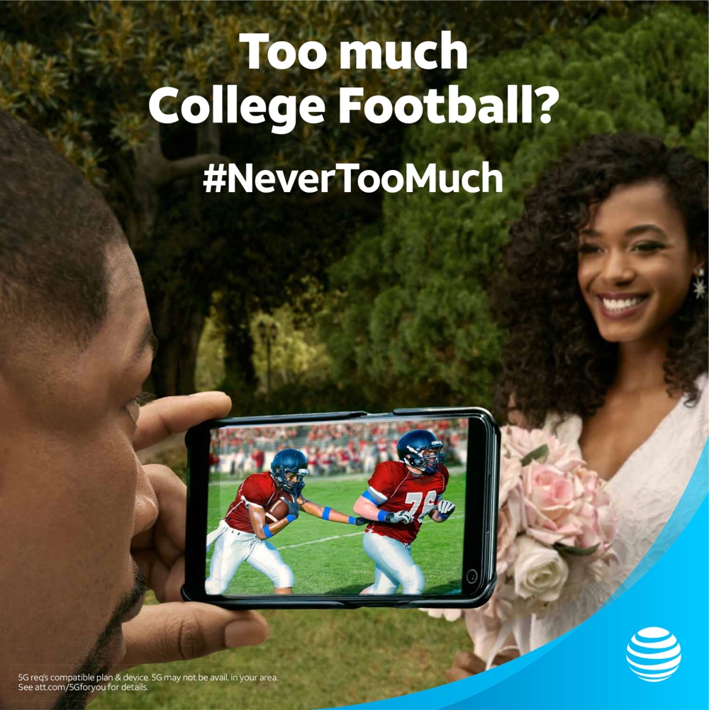 Man holds smartphone displaying football game while a woman in a white dress holding flowers smiles in a green outdoor setting.