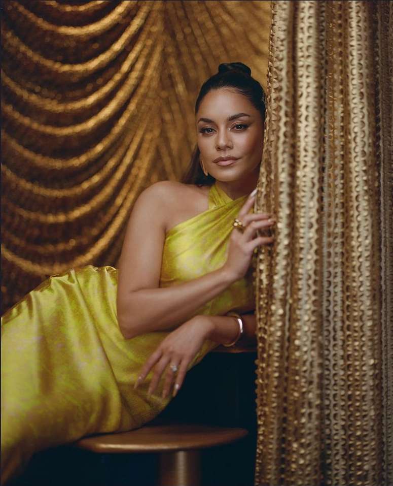 Portrait of Vanessa Hudgens in a yellow dress, gold jewelry, and a hair bun, posing against a textured gold curtain.