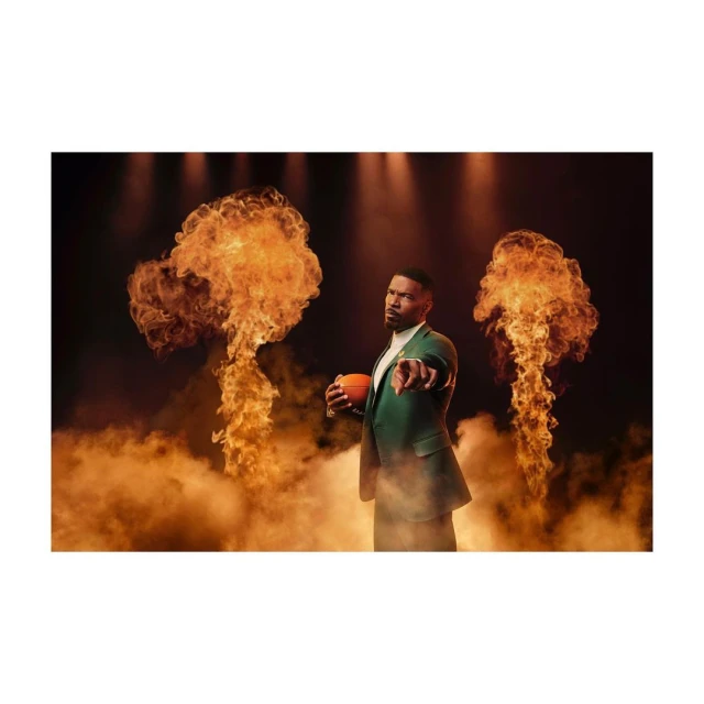 Adult male in a green suit holding a football with finger pointed towards the camera standing in front of flames and smoke.