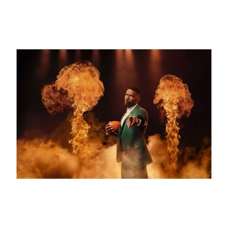Adult male in a green suit holding a football with finger pointed towards the camera standing in front of flames and smoke.