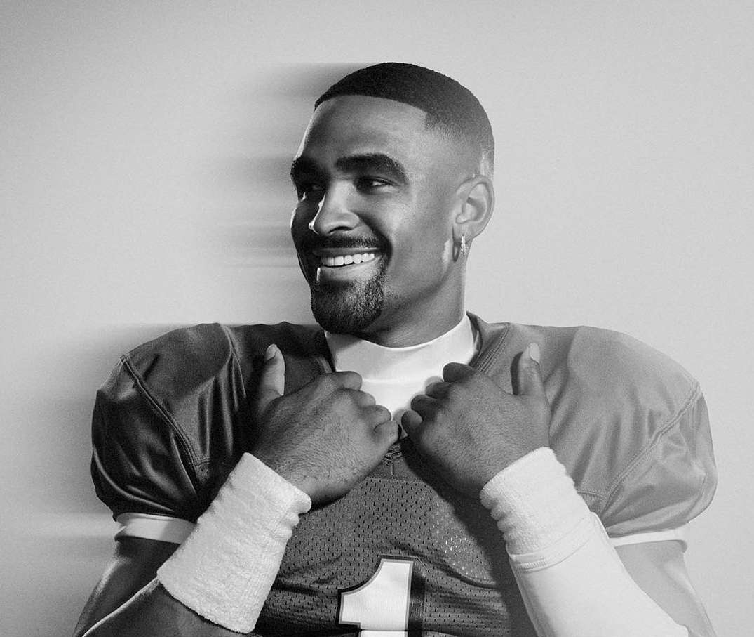 Black and white portrait of a smiling Black male athlete wearing a football jersey with hands clasped. Indoors against a plain background.
