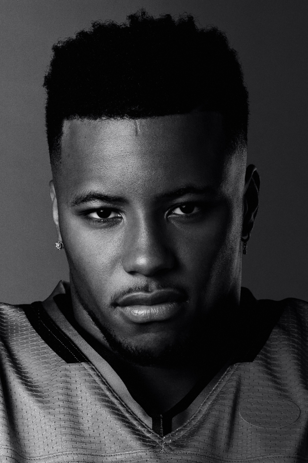 Headshot of a young adult male of African descent wearing a yellow football jersey, set against a dark grey background, captured in black and white.