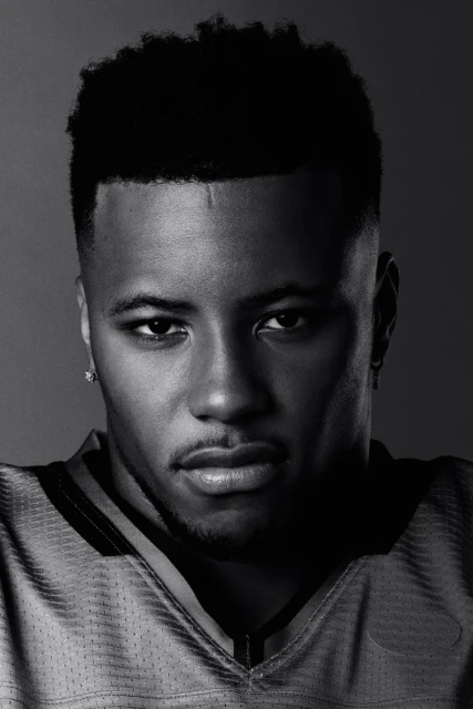 Headshot of a young adult male of African descent wearing a yellow football jersey, set against a dark grey background, captured in black and white.