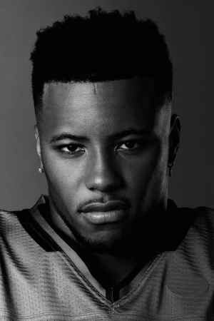 Headshot of a young adult male of African descent wearing a yellow football jersey, set against a dark grey background, captured in black and white.