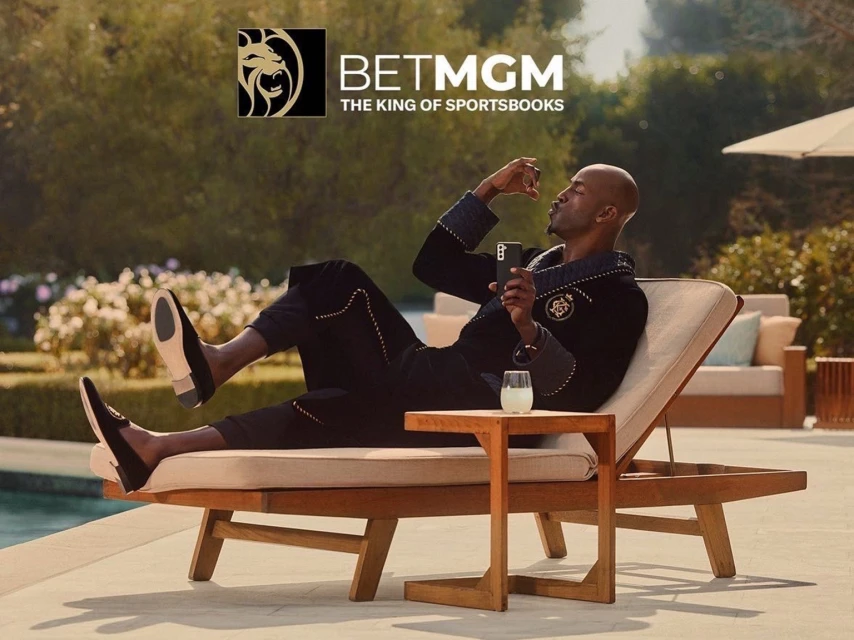 Man lounges poolside in robe, sipping drink and using phone, near BETMGM logo; lush greenery, modern leisure, promoting relaxation.