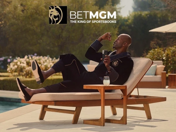 Man lounges poolside in robe, sipping drink and using phone, near BETMGM logo; lush greenery, modern leisure, promoting relaxation.