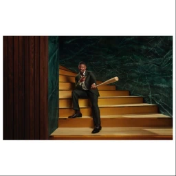Man in a dark suit holding a baseball bat while seated on wooden steps against a green marble wall