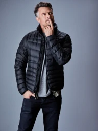 A man in a black puffer jacket and grey t-shirt posing thoughtfully against a neutral grey studio background