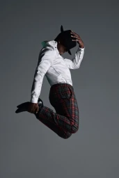 Black man in a white shirt and plaid trousers jumping mid-air while adjusting a black fedora against a plain grey background