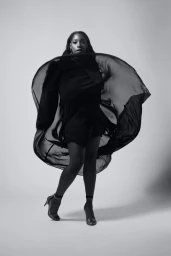 Black and white portrait of a black woman in a flowing sheer cape and heels against a seamless studio background