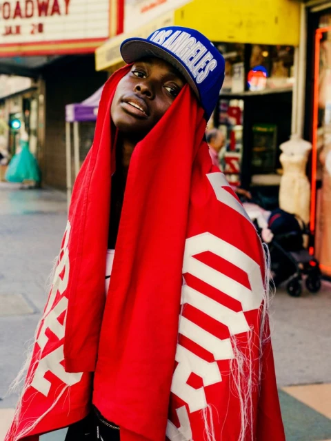 A Black woman wearing a blue baseball cap and a red and white patterned wrap poses in front of a storefront on a city street.