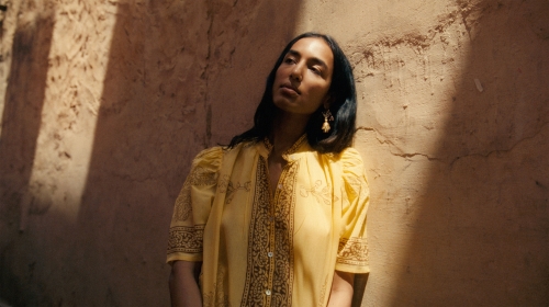 A woman with long dark hair poses in a yellow blouse against a textured brown wall in natural light, showcasing fashion and style.
