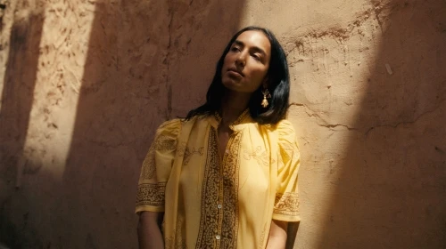 A woman with long dark hair poses in a yellow blouse against a textured brown wall in natural light, showcasing fashion and style.