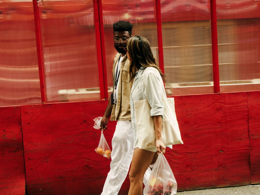 A stylish couple walks together outdoors, holding shopping bags, against a colorful red and white facade with a bright, daytime setting.