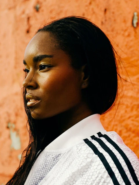 Profile shot of a Black woman wearing a white Adidas track jacket with black stripes, against a textured orange wall background.