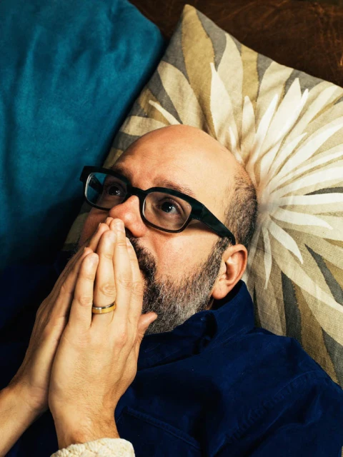Portrait of a bald adult male wearing glasses and a blue shirt, lying on pillows, with hands clasped near his face in a neutral setting.