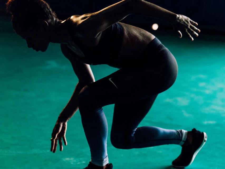 Silhouette of an African American female athlete in a crouched running start position on a green floor with dark lighting.