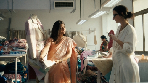 Two women in a clothing workshop. One is wearing a sari and holding fabric, standing next to a dress form with a jacket on it.