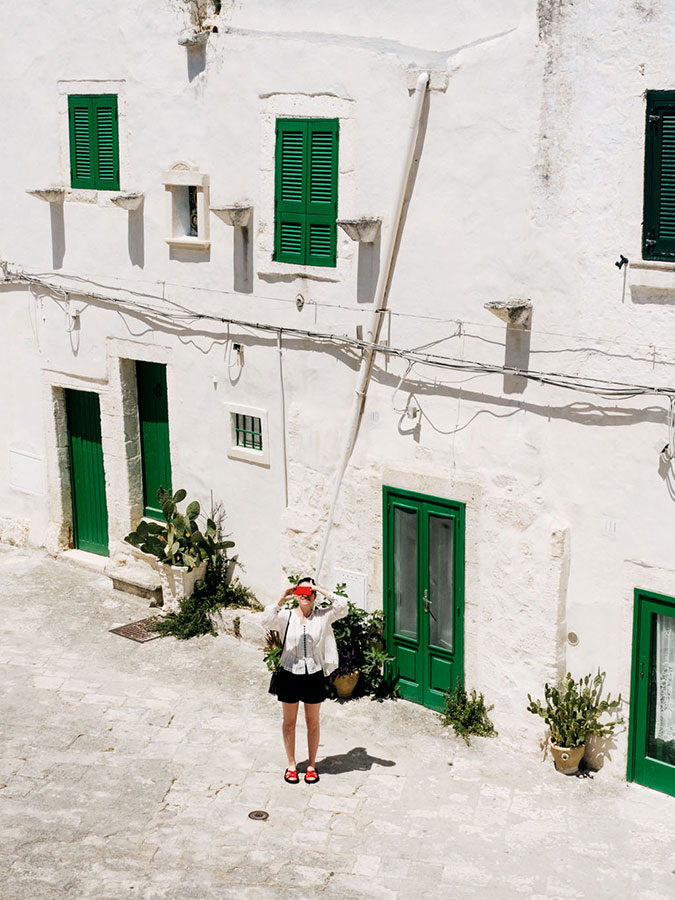 A woman in a white blouse, black shorts, and red visor stands in a courtyard with white buildings and green doors and shutters on a sunny day.