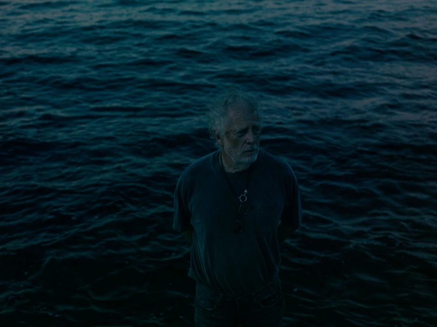 An elderly adult male stands in dark blue water, facing forward with a serious facial expression in an outdoor setting with natural light.