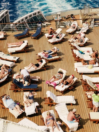 Aerial view of numerous people relaxing in lounge chairs on the deck of a cruise ship, with blue ocean waves in the background.