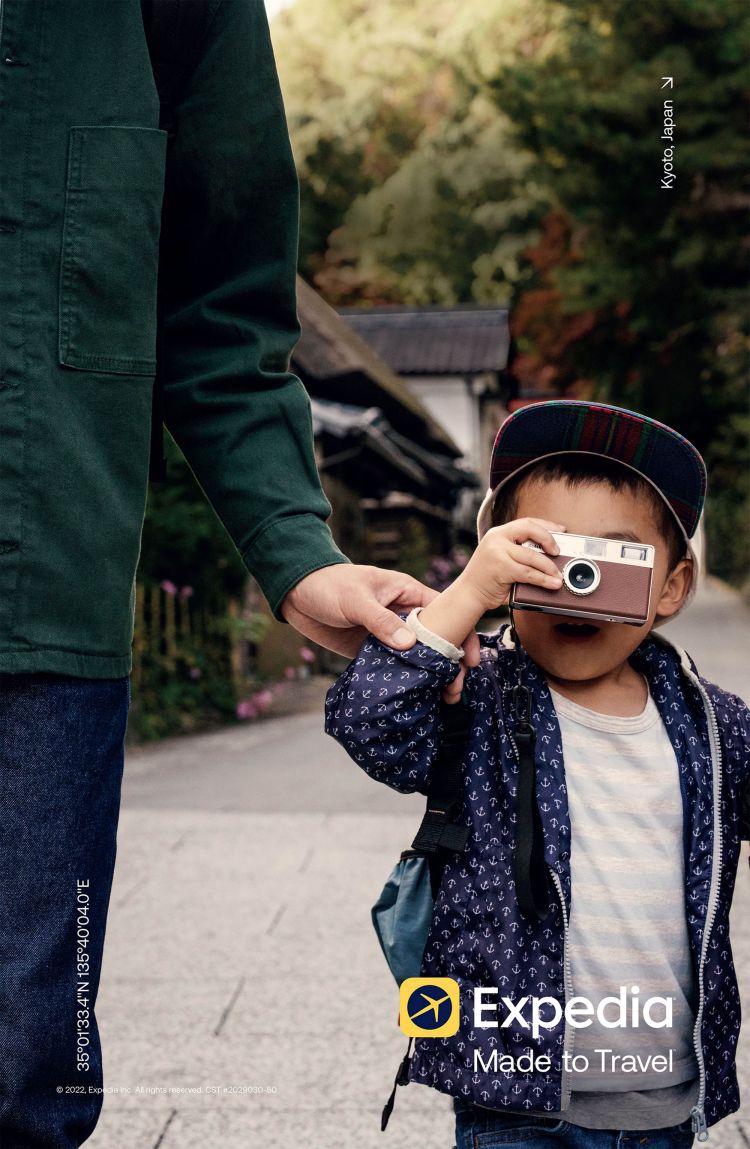 Child holding a camera with adult's hand on sidewalk in Kyoto, Japan. Travel scene features architecture and nature. Expedia logo visible.
