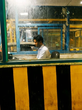 Man sits in a toll booth with yellow and black striped barrier in view. The toll booth has dirty windows and a blue frame.