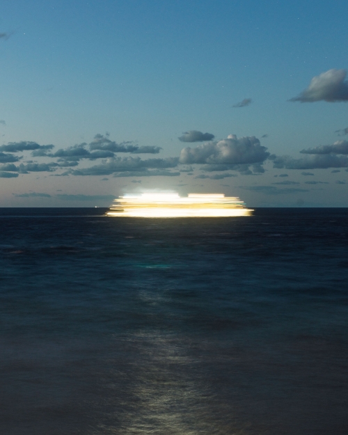 Long exposure of a boat at sea creating light trails, under a twilight sky with scattered clouds, ocean reflecting light.