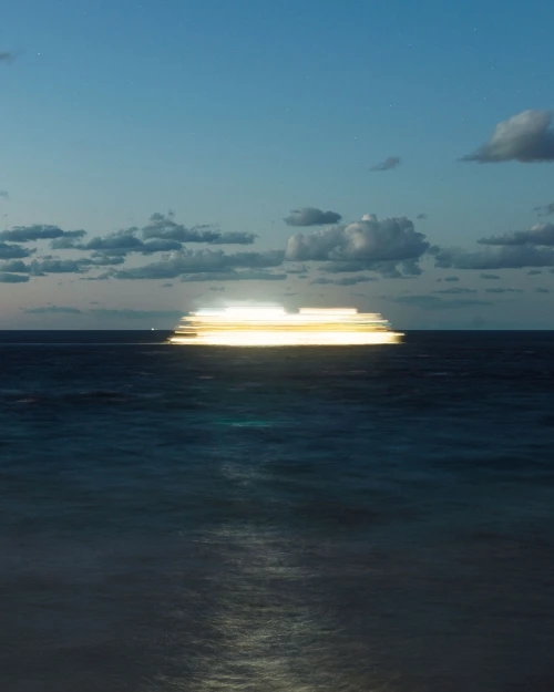 Long exposure of a boat at sea creating light trails, under a twilight sky with scattered clouds, ocean reflecting light.