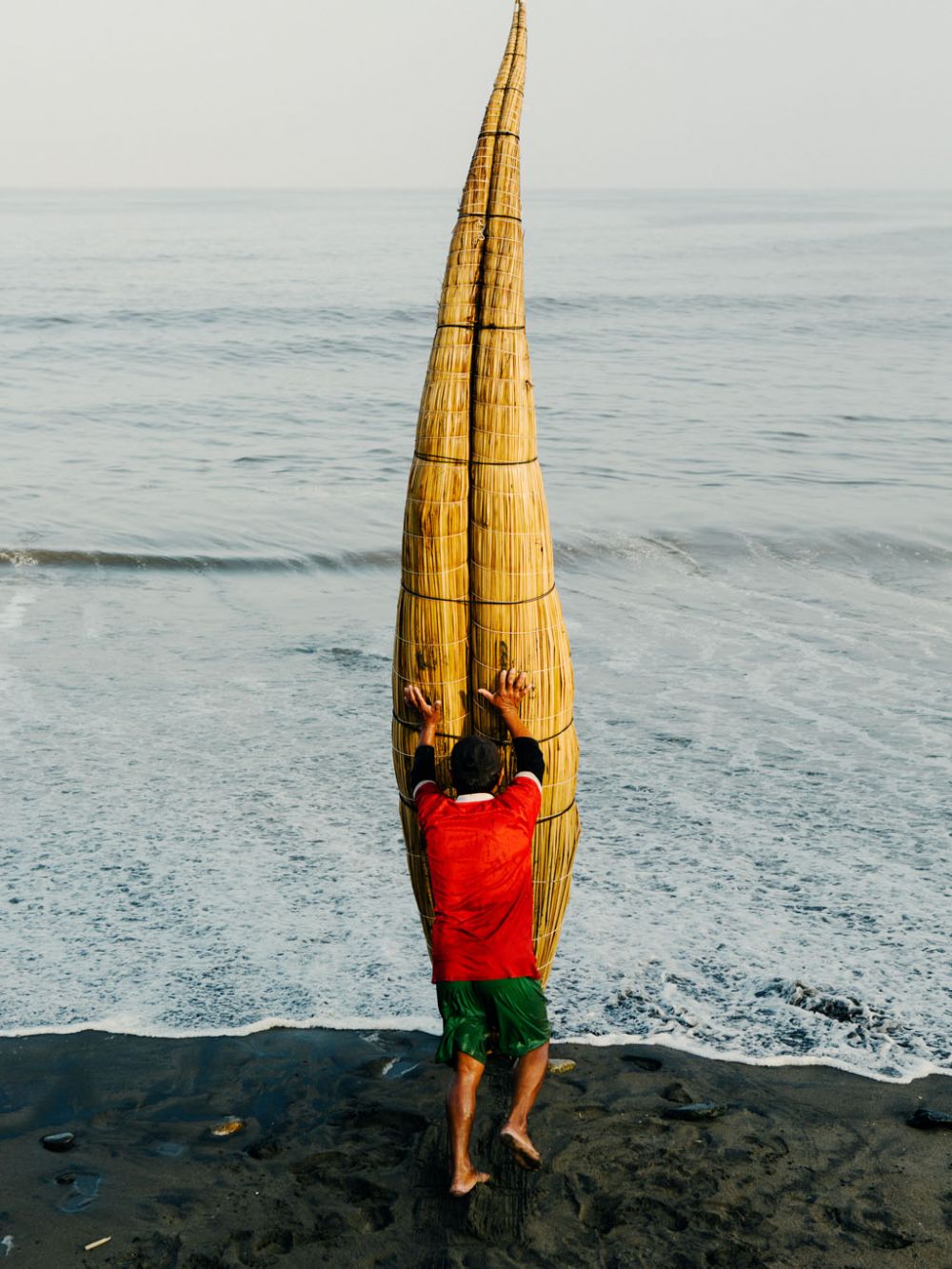 A man in a red shirt and green shorts stands at the edge of the ocean with a traditional reed boat along the beach shore.