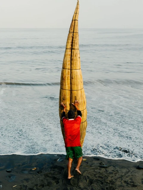 A man in a red shirt and green shorts stands at the edge of the ocean with a traditional reed boat along the beach shore.