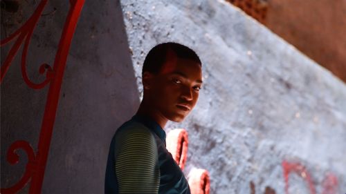 An adult with short hair poses with a slight smile near an aged cement wall and bright red iron bars on the left side of the frame.