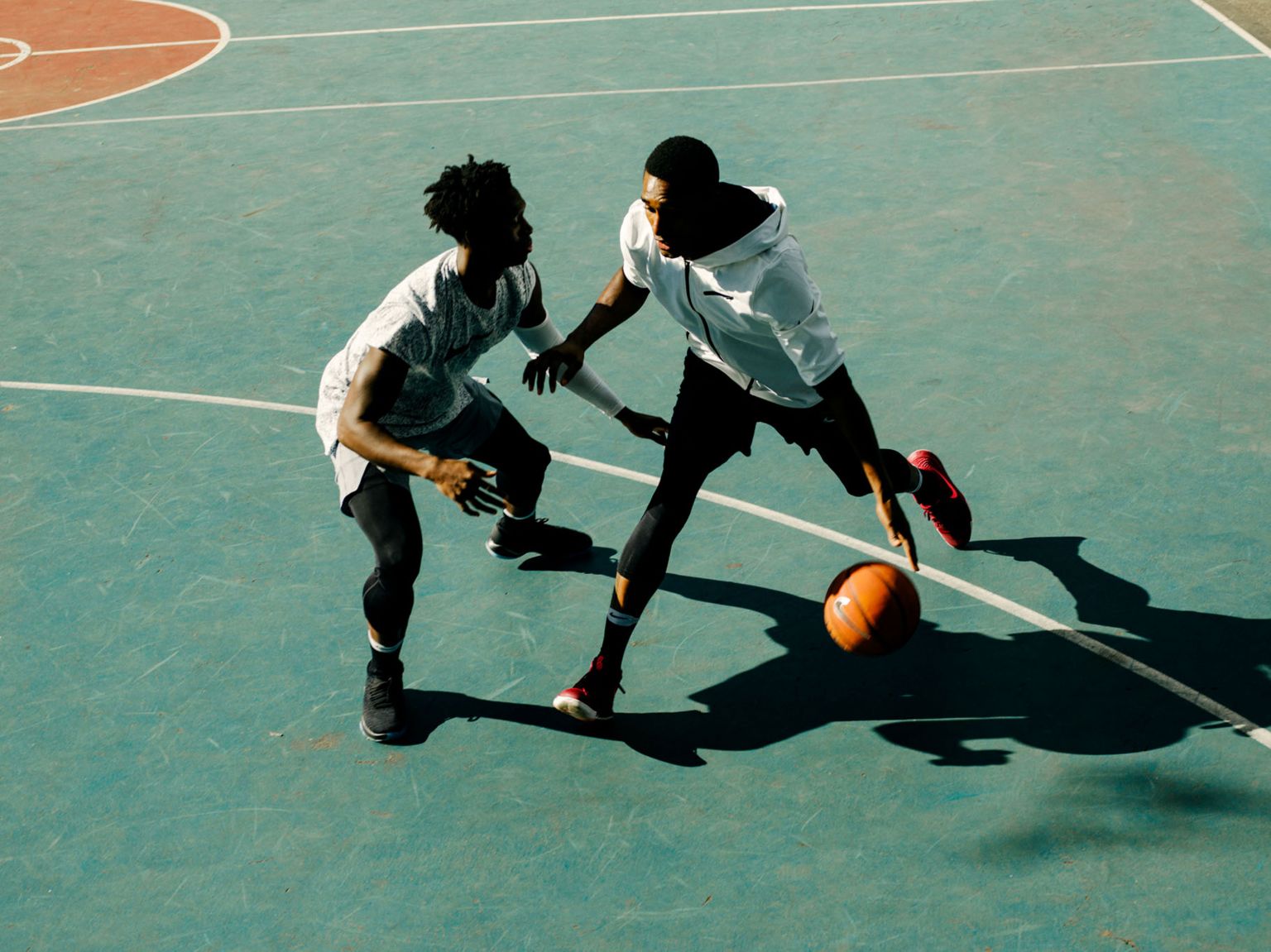Two African-American men playing basketball on an outdoor court, captured in a high-angle shot during the daytime with clear shadows.