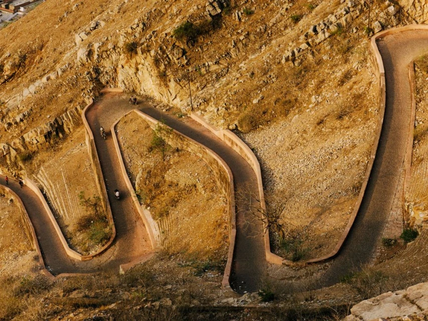 Aerial view of a winding mountain road with rocky terrain, showcasing a challenging route through arid, natural landscape and sunlight.